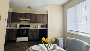 Un comedor con una mesa redonda dispuesta para dos personas, un ventilador de techo, armarios de cocina y un ventanal con persianas.