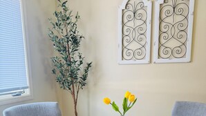 Un comedor con una mesa redonda, dos sillas grises, un jarrón con flores amarillas y paneles metálicos decorativos en la pared.