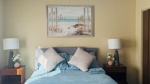 Un dormitorio con cama, ventilador de techo, pintura de playa, dos mesitas de noche con lámparas y una mesita de noche con un jarrón de flores.