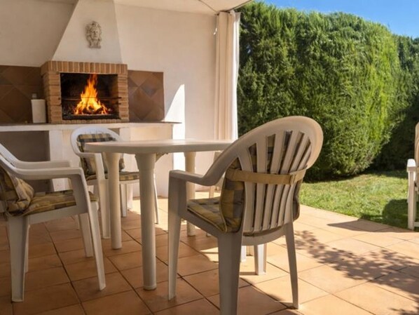 Eine Terrasse im Freien mit Kamin aus Backstein, weißen Stühlen und einem ordentlich geschnittenen Heckenstreifen.
