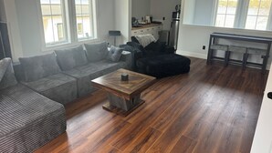 A living room with a sectional sofa, a wooden coffee table, and a kitchen area with a refrigerator and a coffee maker.