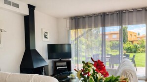 A living room with a TV, a black fireplace, a glass table, and a view of a garden. - Family home 50m from the beach - 8 guests (Palamós)