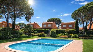 A swimming pool with a tiled deck, surrounded by well-maintained lawns and trees, with buildings in the background. - Family home 50m from the beach - 8 guests (Palamós)