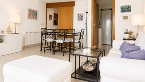 Uma sala de estar moderna com uma mesa de jantar de vidro, sofás brancos e uma cozinha visível por uma porta aberta.