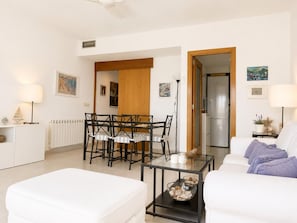 A modern living room with a glass dining table, white sofas, and a kitchen area visible through an open door. - Apartment with sea view in La Fosca - details coming soon (Palamós)