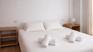 Une chambre d’hôtel minimaliste comprenant un lit, deux oreillers, une table de chevet avec une lampe et une étagère en bois.