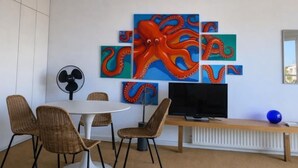 Una habitación con pared blanca, una mesa redonda con sillas de mimbre, un televisor de pantalla plana sobre un soporte de madera y una obra de arte de un pulpo grande.