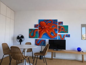 A room with a white wall, a round table with wicker chairs, a flat-screen TV on a wooden stand, and a large octopus artwork. - Studio sea view in La Fosca - 2 guests (Palamós)