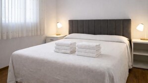 Een keurig opgemaakt bed met witte lakens, twee bedlampjes en een raam met dunne gordijnen.