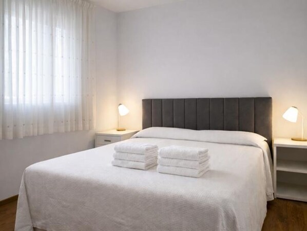 A neatly made bed with white linens, two bedside lamps, and a window with sheer curtains.