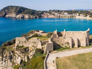 A coastal fortification with a paved pathway leading to it, surrounded by greenery and overlooking a clear blue sea.