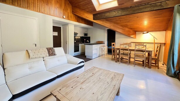 Una habitación estilo loft con techo de madera, un sofá blanco, una mesa de comedor de madera y una cocina al fondo.