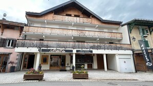 Un edificio de dos pisos con fachada de madera, un balcón con barandilla metálica y locales comerciales en la planta baja.