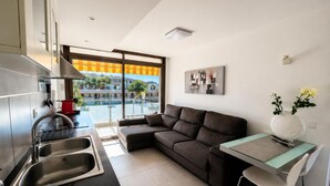 A modern kitchen with a built-in oven, a dining area with a glass table and chairs, a sofa, and a large window with a view of a pool and buildings. - Comfortable apartment near the beach - 4 guests (Palamós)