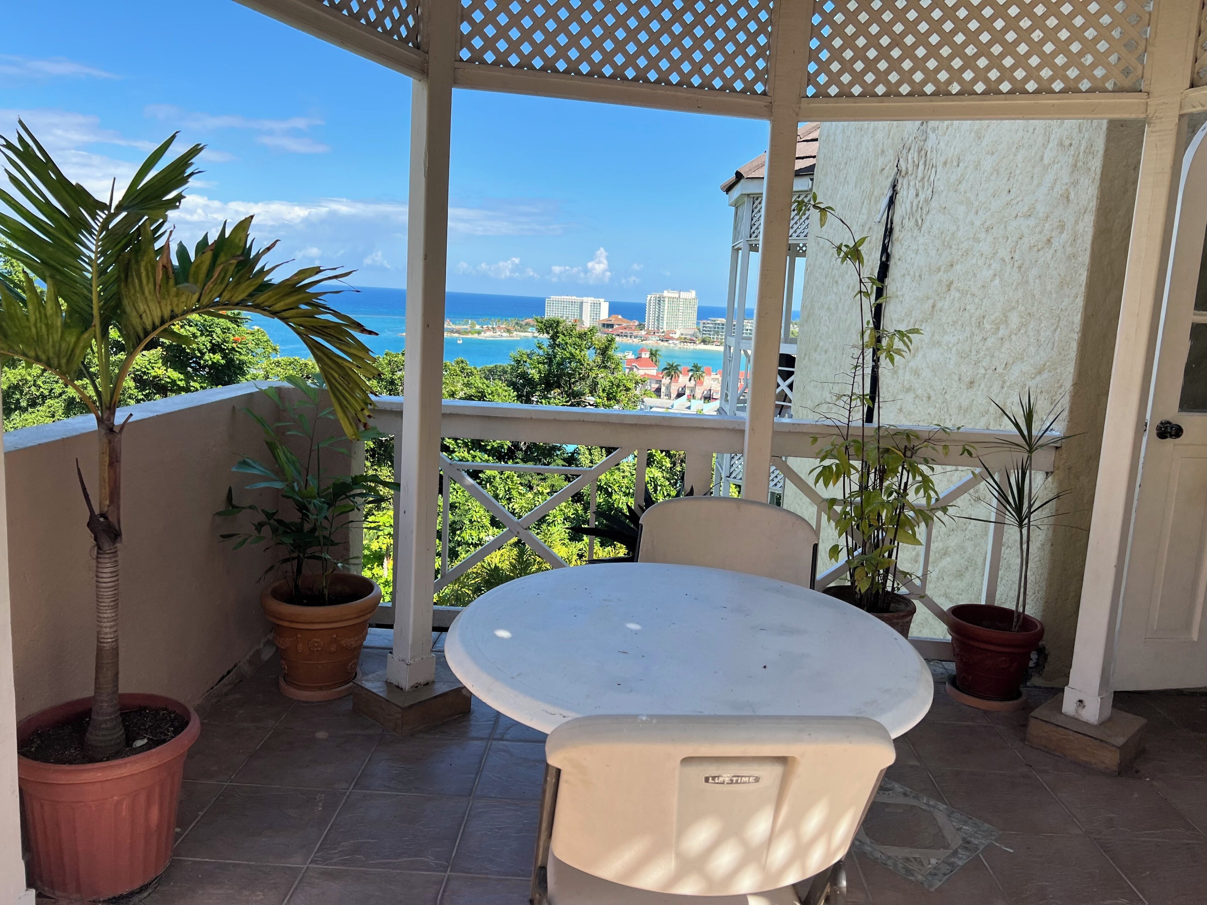 Ein überdachter Außenbereich mit Tisch und Stühlen, Blumentöpfen und Blick auf das Meer und die Gebäude.