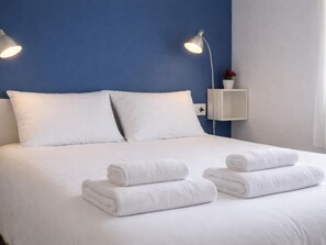 A neatly made bed with white linens and two rolled towels, a bedside table with a lamp, and a small shelf with a flower. - Renovated apartment, near beach - 4 guests (Palamós)