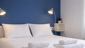 A neatly made bed with white linens and two rolled towels, a bedside table with a lamp, and a small shelf with a flower. - Renovated apartment, near beach - 4 guests (Palamós)