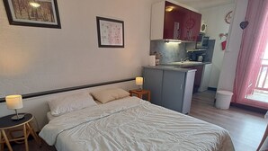 Un dormitorio con cama, mesitas de noche, un taburete pequeño, una kitchenette con microondas y un cubo de basura.