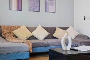 Ein modernes Wohnzimmer mit einem grauen Sofa, beigen und weißen Kissen, einer blauen Decke und einer Holzskulptur auf einem dunklen Couchtisch.