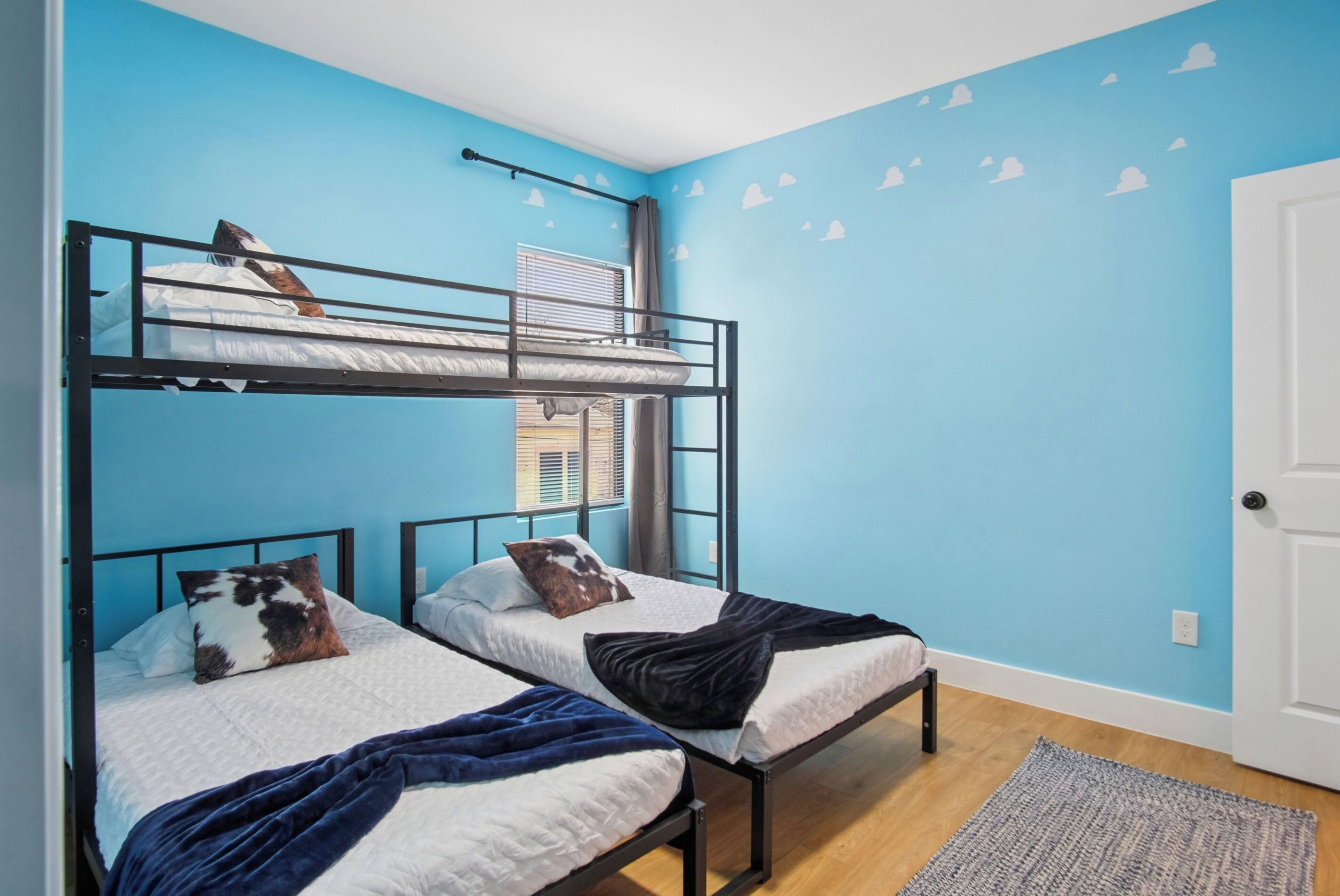 A room with two bunk beds, a window with blinds, a door, and a wall with cloud decals.