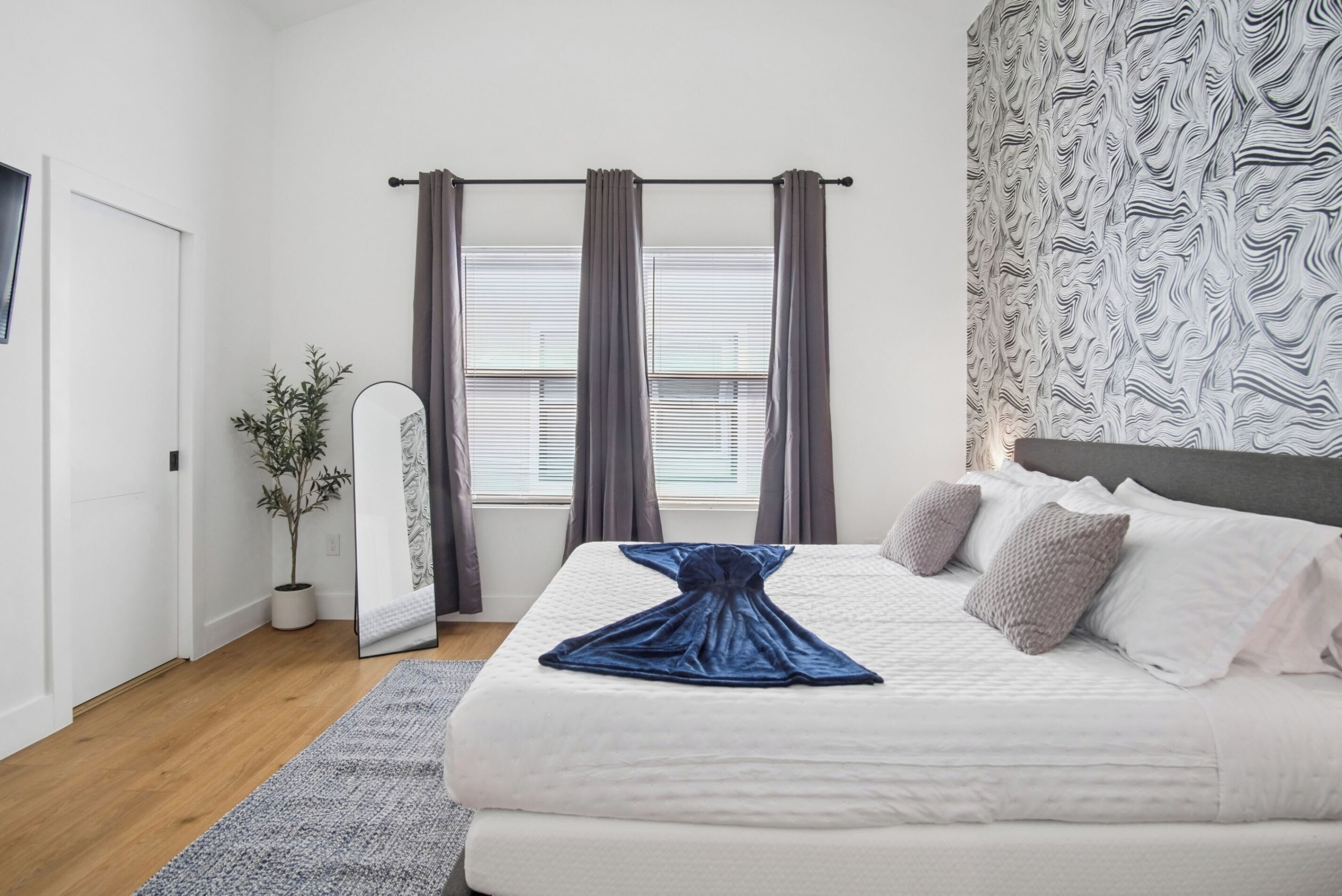Een slaapkamer met een bed, gordijnen, een spiegel, een plant en een behang met patroon.