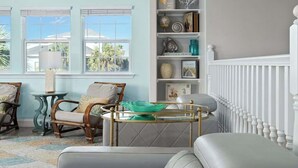 A room with a view of palm trees, a white railing, and a bookshelf with various items. - Port A Beach House | Pool, Game Room and Patio! (Port Aransas)