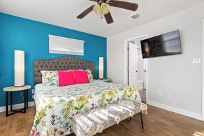 Ein Schlafzimmer mit einem gepolsterten Kopfteil, einem blumigen Bettbezug, einem Deckenventilator, einem Fernseher und einem Fenster mit Jalousien.