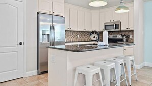A kitchen with white cabinets, a stainless steel refrigerator, a microwave, a sink, and a bar stool. - Port A Beach House | Pool, Game Room and Patio! (Port Aransas)