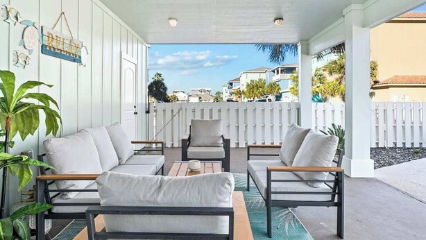 A covered outdoor seating area with white walls, a wooden table, and a white fence in the background. - Port A Beach House | Pool, Game Room and Patio! (Port Aransas)