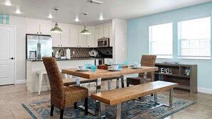 A modern kitchen with white cabinets, a wooden dining table, wicker chairs, and a bench. - Port A Beach House | Pool, Game Room and Patio! (Port Aransas)