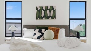 Ein Hotelzimmer mit einem großen Bett, zwei Nachttischlampen, einem an der Wand befestigten abstrakten Kunstwerk und Blick durch die Fenster.