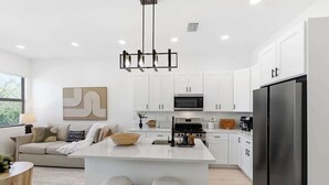 Une cuisine moderne avec des meubles blancs, un îlot central doté de tabourets, un réfrigérateur en acier inoxydable et un luminaire suspendu.