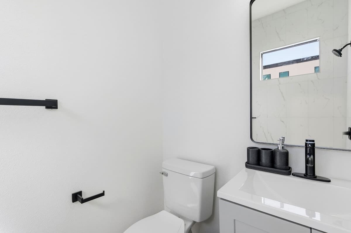 A modern bathroom with a white toilet, a black towel rack, a black soap dispenser, and a mirror with a view of a building.
