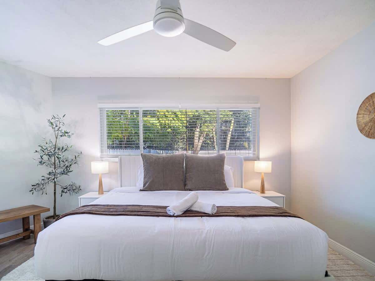 A bedroom with a bed, two bedside lamps, a ceiling fan, a window with blinds, a bench, and a potted plant.