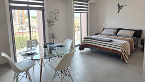 A modern bedroom with a bed, a glass dining table with chairs, and large windows with blinds. - LUMEN - Loft Modern Downtown GDL | Gym & Coworking (Guadalajara)