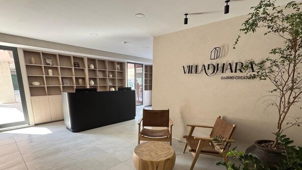 A reception area with a reception desk, two wooden chairs, a round wooden ottoman, and a potted plant. - LUMEN - Loft Modern Downtown GDL | Gym & Coworking (Guadalajara)