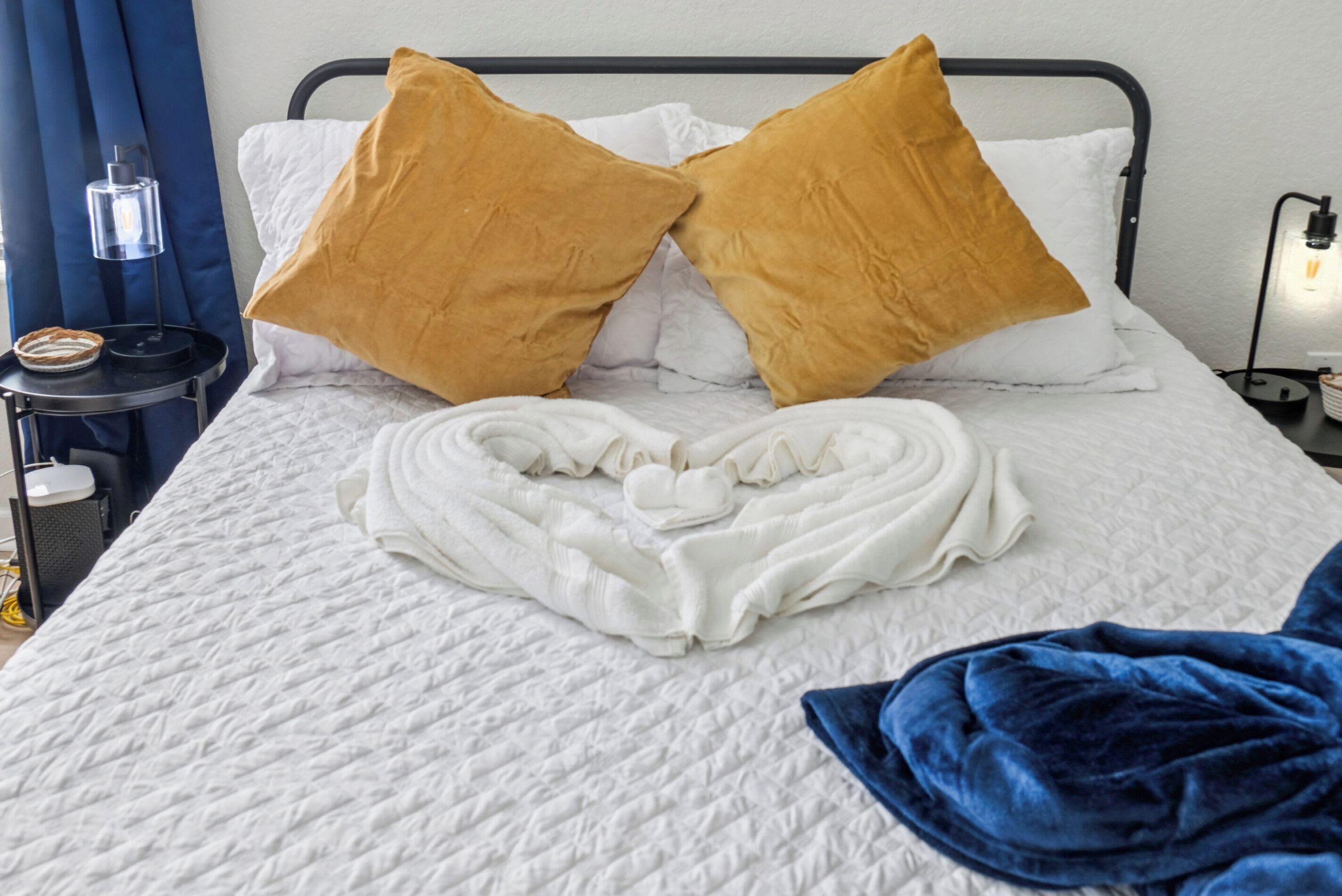 Een net opgemaakt bed met twee kussens, een handdoek in de vorm van een hart en een blauwe deken.