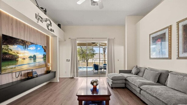 Una sala de estar moderna con un televisor de pantalla plana grande, un sofá seccional gris, una mesa de centro de madera y una puerta de cristal que da a un área exterior con piscina.