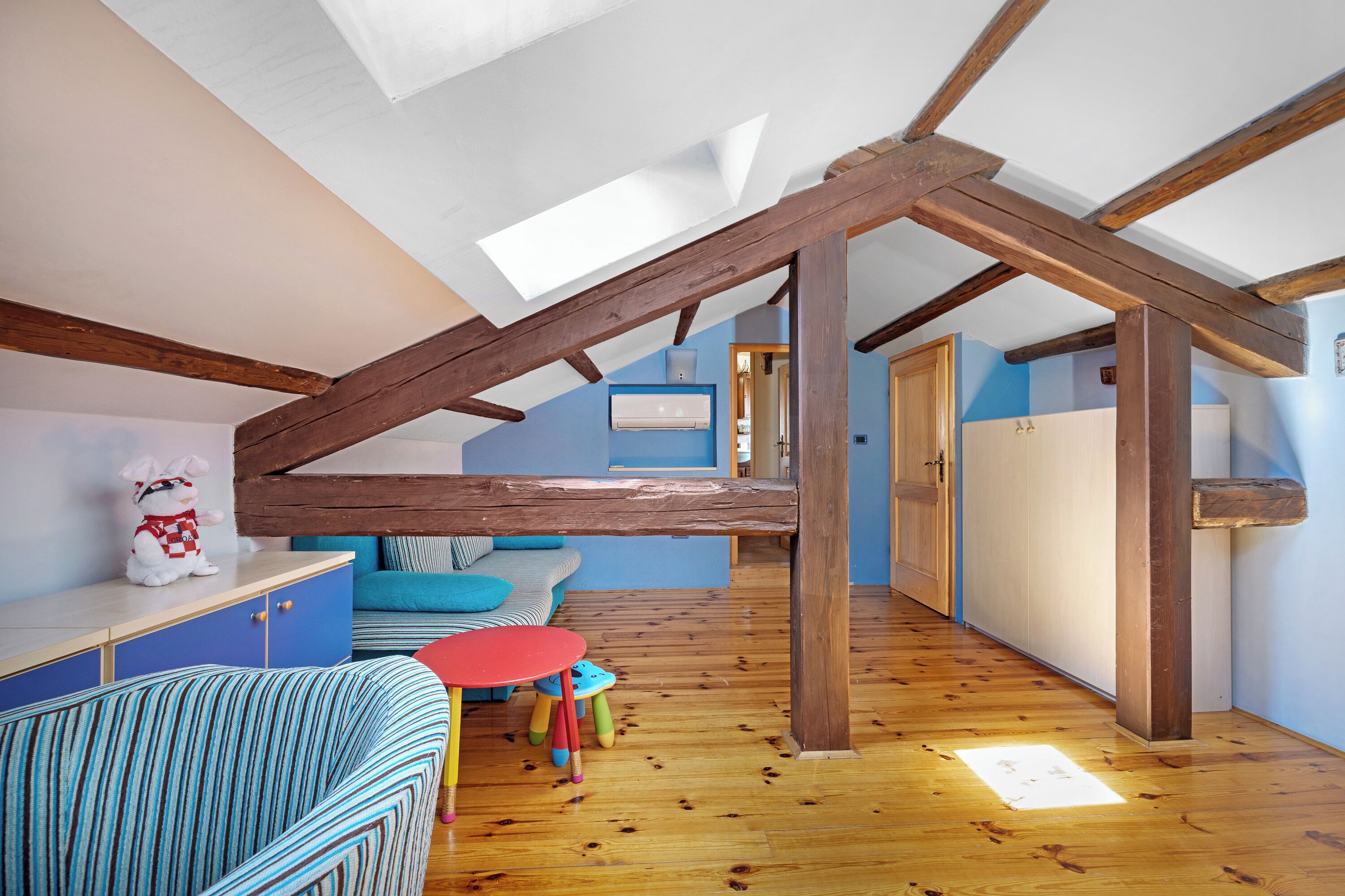 Dormitorio estilo loft con techo inclinado, vigas de madera, tragaluces, un sofá azul, una mesa roja y un taburete pequeño.