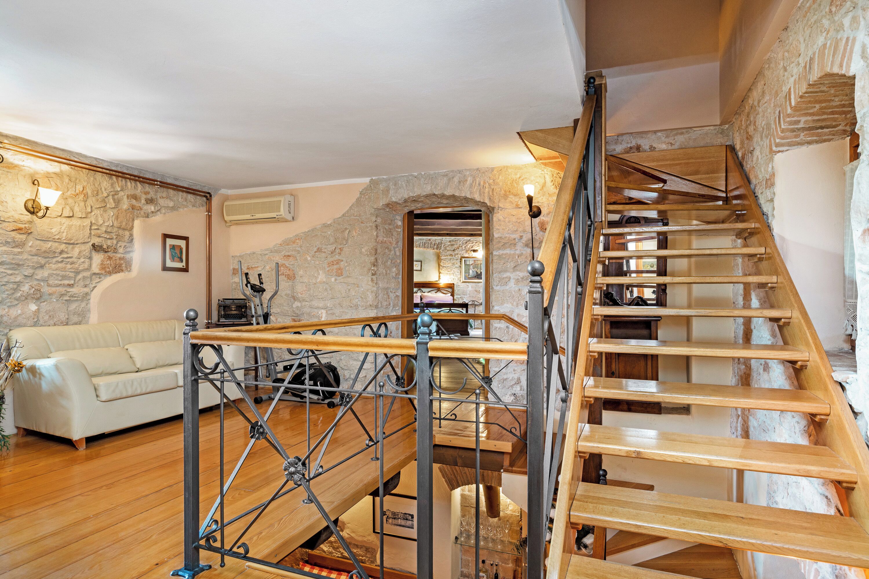 Un escalier avec des marches en bois et des rampes en métal, un canapé blanc et un mur en pierre dans une pièce dont le sol est recouvert de parquet.