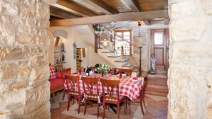Un comedor con mantel a cuadros, sillas de madera, una escalera y paredes de piedra.