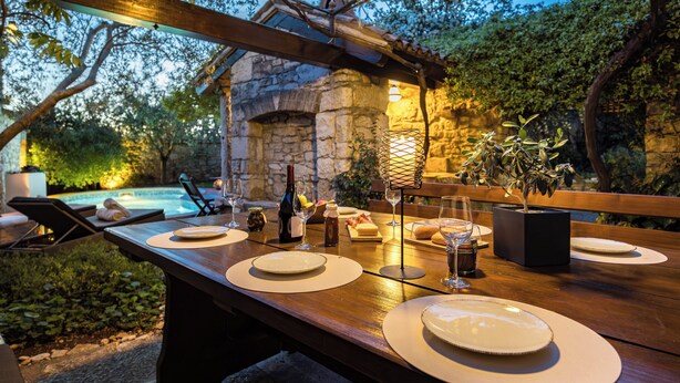 Un área de comedor al aire libre con pared de piedra, pérgola de madera y piscina al fondo.
