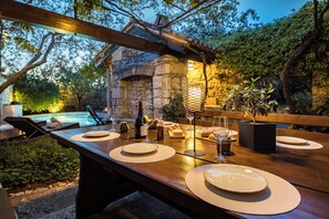 Un área de comedor al aire libre con pared de piedra, pérgola de madera y piscina al fondo.