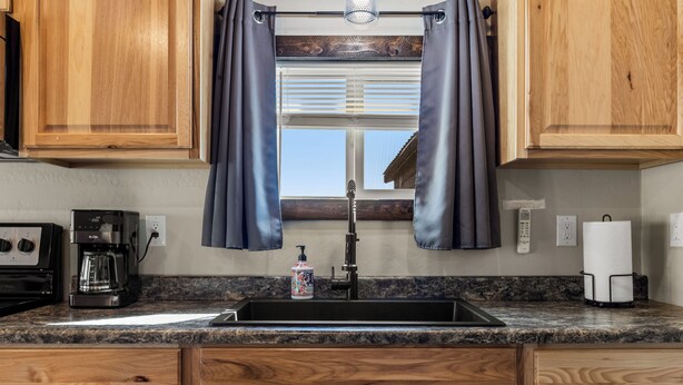 A kitchen with wooden cabinets, a stainless steel sink, a coffee maker, and a window with blinds. - The Roost Cabins LLC #5 Near Glacier National Park (Columbia Falls)