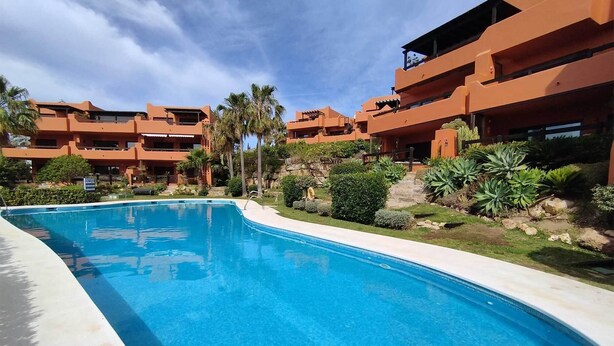 A swimming pool surrounded by multi-story buildings with balconies, lush greenery, and a clear sky. - New Galera Gardens (Estepona)