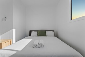 Ein minimalistisch eingerichtetes Hotelzimmer mit einem ordentlich bezogenen Bett, einem kleinen Nachttisch und einem Fenster mit Blick zum Himmel.