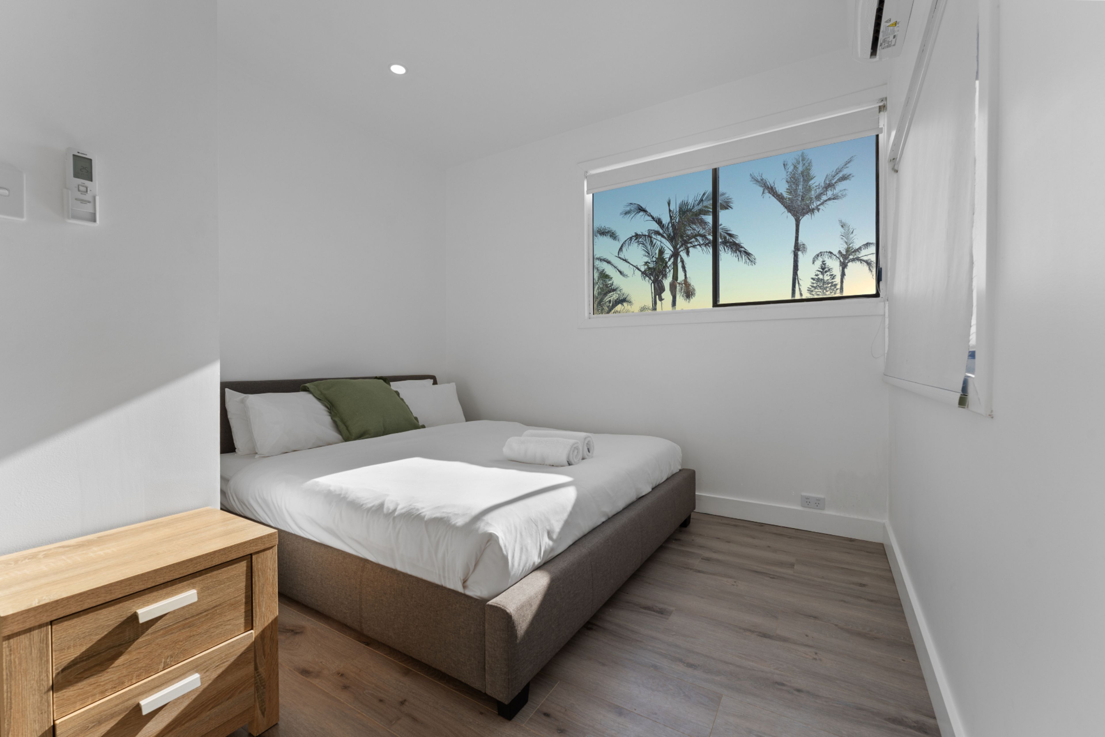 A bedroom with a bed, a wooden nightstand, a window with a view of palm trees, and a wall-mounted thermostat.