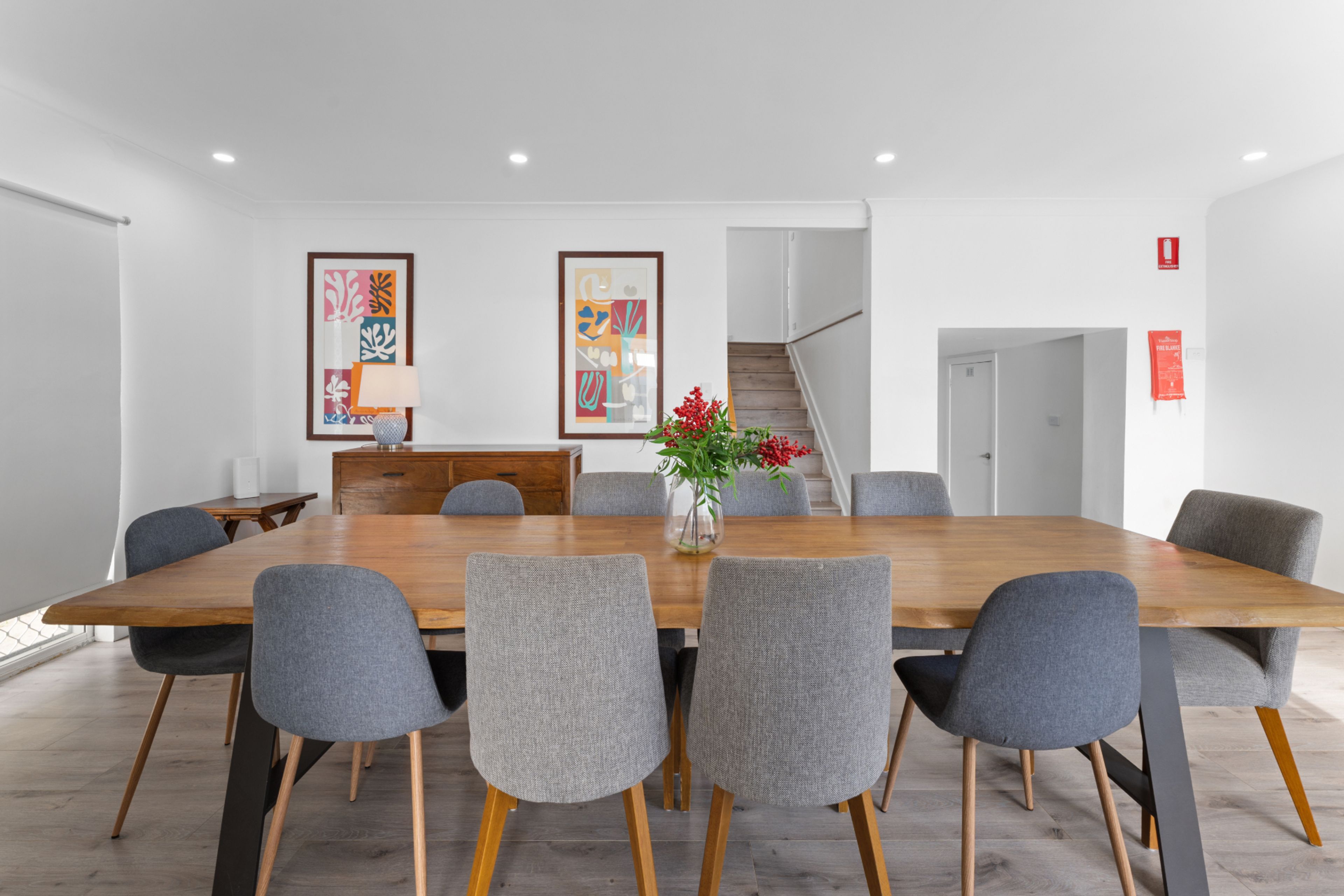 Une salle de conférence avec une longue table en bois, six chaises grises rembourrées, deux tableaux abstraits encadrés accrochés au mur, un vase avec des fleurs rouges sur la table et un escalier en arrière-plan.