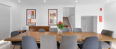 A conference room with a long wooden table, six grey upholstered chairs, two framed abstract paintings on the wall, a vase with red flowers on the table, and a staircase in the background.