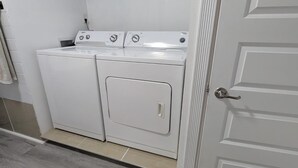 Une buanderie équipée de deux machines à laver et deux sèche-linge à chargement frontal, tous deux de couleur blanche, avec une porte et un sol carrelé.